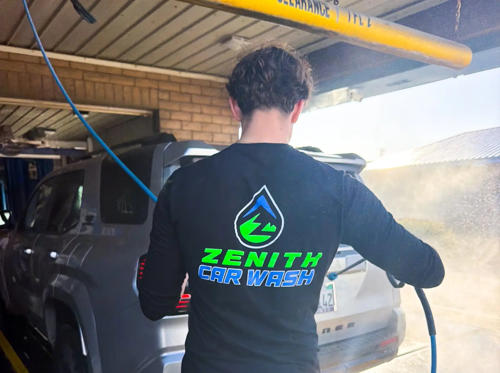 A Zenith crew member in a Zenith Car Wash shirt pre-washing an SUV by hand with a pressure wand at the tunnel entrance